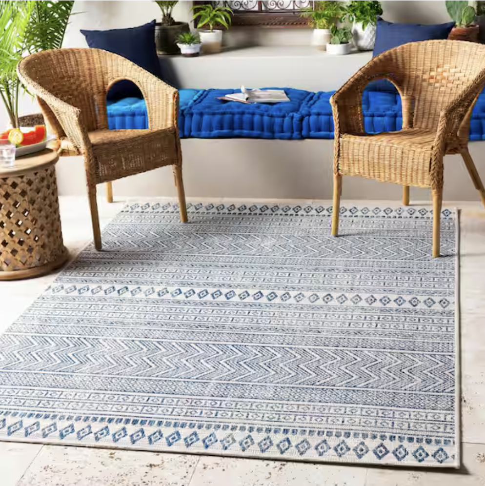 Rattan chairs sit on a patterned blue and white rug alongside a small wooden side table. A cushioned bench with blue pillows and various plants accents the background.