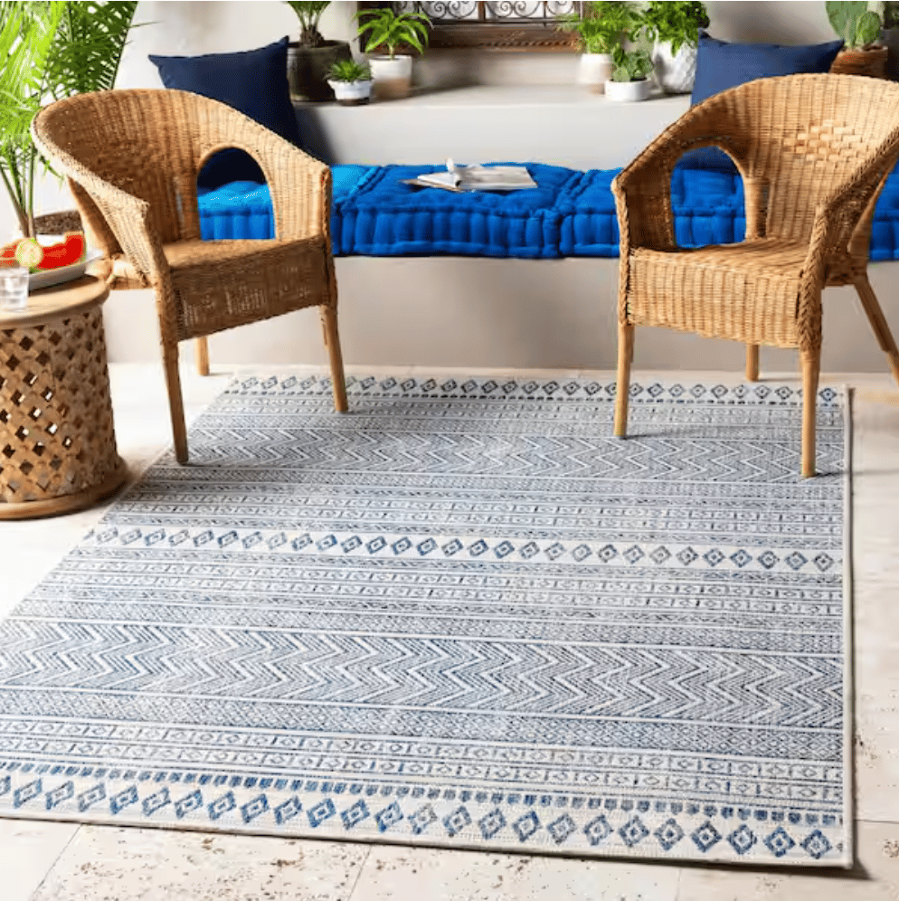 Rattan chairs sit on a patterned blue and white rug alongside a small wooden side table. A cushioned bench with blue pillows and various plants accents the background.