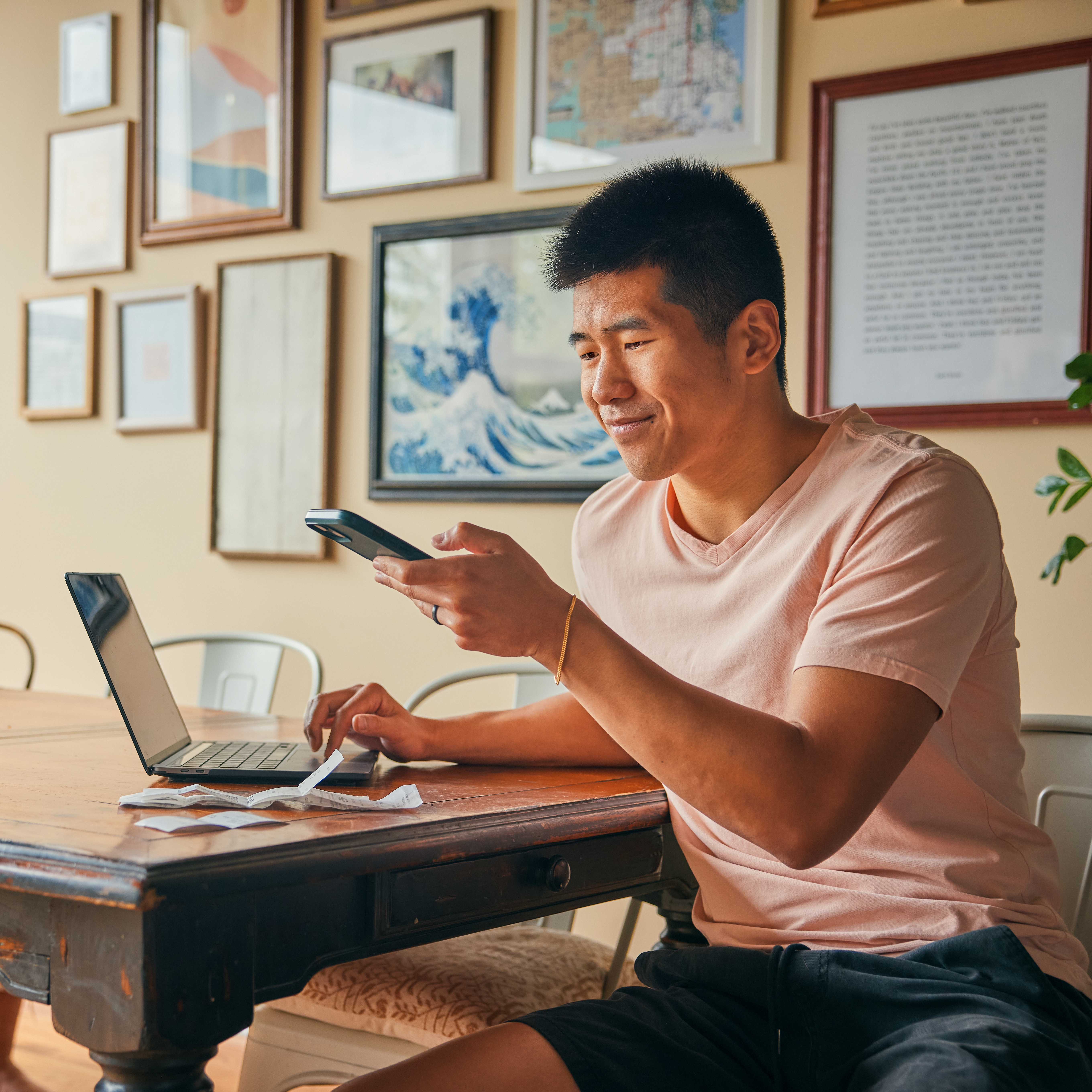 A person in a pink shirt is using a smartphone and a laptop at a wooden table. Various framed artworks are on the wall behind them.