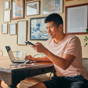 A person in a pink shirt is using a smartphone and a laptop at a wooden table. Various framed artworks are on the wall behind them.