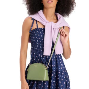 The Kate Spade Leather Dome Crossbody is a small, green bag featuring a curved shape, a gold-tone chain strap, and a zip closure. It is worn over a shoulder in the image.