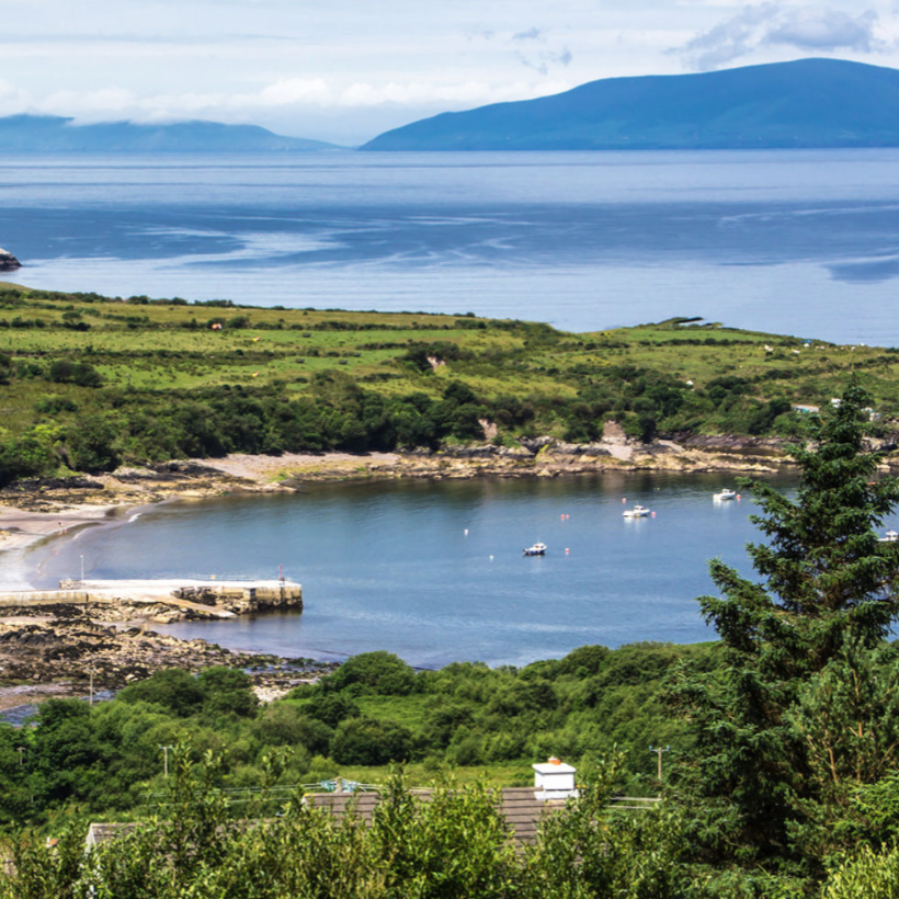Scenic coastal landscape featuring a small bay with a sandy beach, rocky shoreline, and a few boats in the water. Green hills and distant islands are visible under a partly cloudy sky.