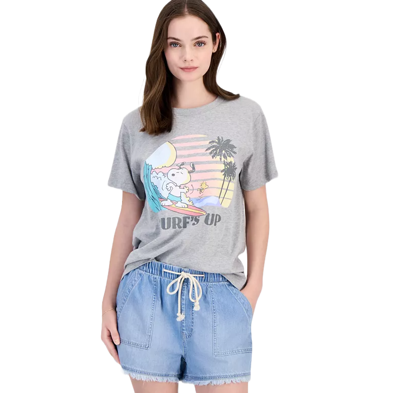 A woman is wearing a gray T-shirt featuring a cartoon character surfing with the text \“Surf's Up\“ and paired with light blue denim shorts.