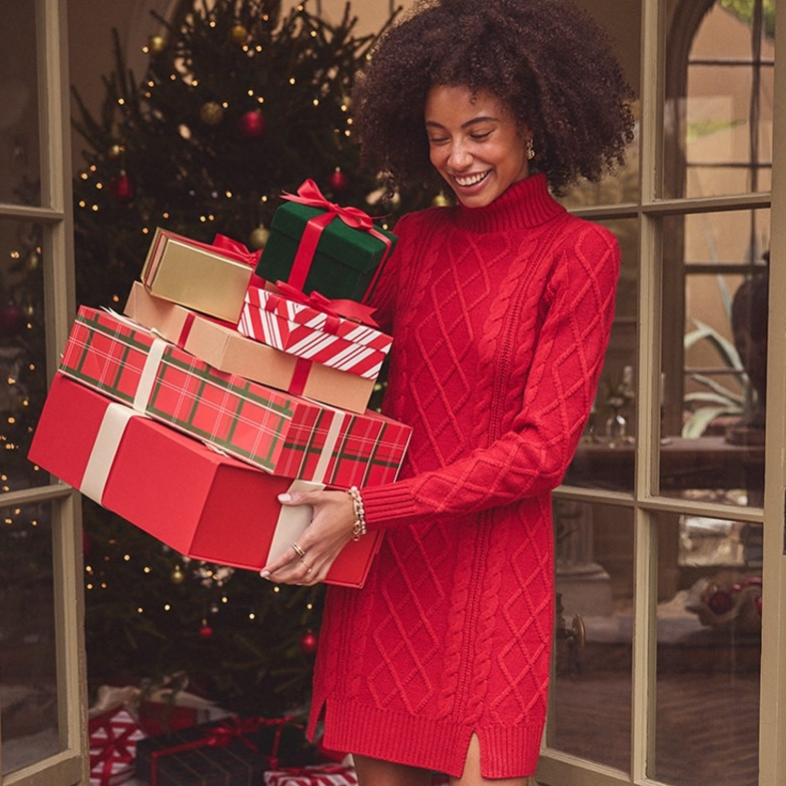 A person in a red cable-knit sweater dress carries stacked gift boxes, accompanied by a Labrador wearing a plaid bandana. A Christmas tree with ornaments is visible in the background.