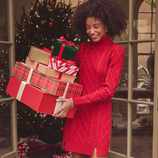 A person in a red cable-knit sweater dress carries stacked gift boxes, accompanied by a Labrador wearing a plaid bandana. A Christmas tree with ornaments is visible in the background.