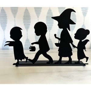This image shows a decorative silhouette cutout of children dressed in various Halloween costumes. They appear to be trick-or-treating, with costumes including a zombie, a kid holding a pumpkin bucket, a witch, and a fairy. This type of decoration is often used for Halloween-themed settings.