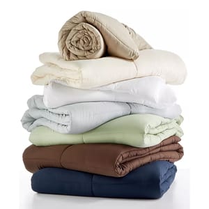 Stacked comforters in various colors, including beige, white, light gray, light green, brown, and navy blue.