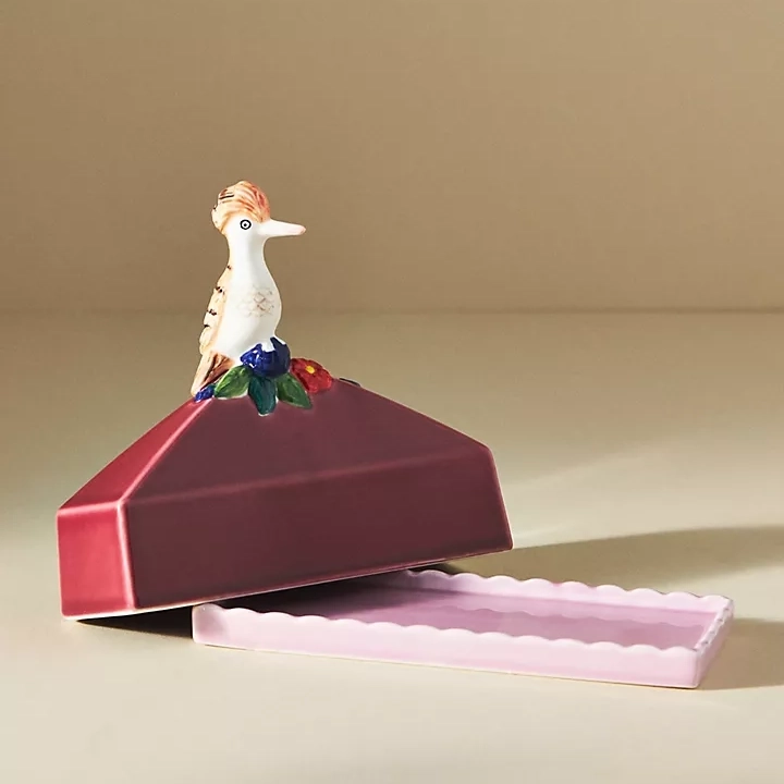 A whimsical bird figurine perched atop an open, maroon jewelry box with a scalloped pale pink base.