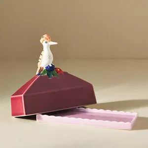 A whimsical bird figurine perched atop an open, maroon jewelry box with a scalloped pale pink base.