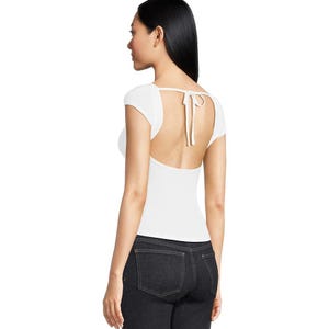 The Free People Tie-Back Tee is a fitted white top with cap sleeves, featuring an open back design tied with thin straps at the neck. It is paired with dark denim jeans.
