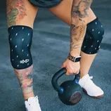 A person with tattoos is exercising with a kettlebell, wearing black knee sleeves with lightning bolt patterns, a smartwatch, and white athletic shoes.