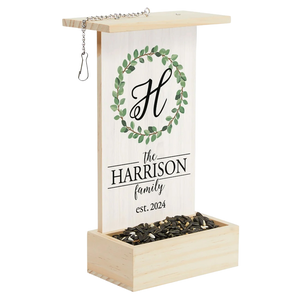 Wooden bird feeder with chain for hanging, featuring a floral wreath and personalized family name with a large initial. The base holds birdseed, and the design includes \“est. 2024\“ text.