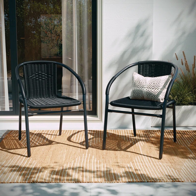 Two black outdoor rattan stacking chairs with curved armrests and a woven design are shown. One chair features a decorative pillow with a geometric pattern. They are placed on a beige outdoor rug near a sliding glass door.