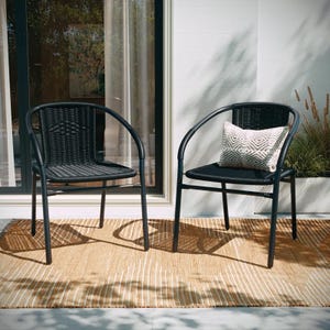 Two black outdoor rattan stacking chairs with curved armrests and a woven design are shown. One chair features a decorative pillow with a geometric pattern. They are placed on a beige outdoor rug near a sliding glass door.