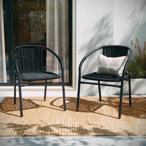 Two black outdoor rattan stacking chairs with curved armrests and a woven design are shown. One chair features a decorative pillow with a geometric pattern. They are placed on a beige outdoor rug near a sliding glass door.