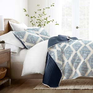A neatly made bed features a quilt with a geometric pattern in blue and beige, placed against a woven headboard. A small plant and a lamp are on a wooden bedside table, and the room is bright with natural light from large windows.