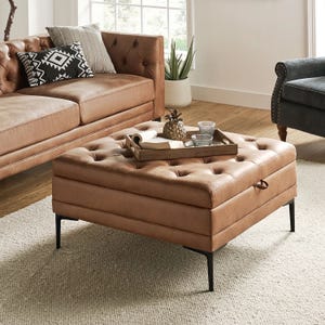 A tan, tufted ottoman with black metal legs serves as a coffee table, adorned with a tray holding decorative items. It's paired with a brown leather sofa, a gray armchair with stud accents, and a neutral area rug on wooden flooring.