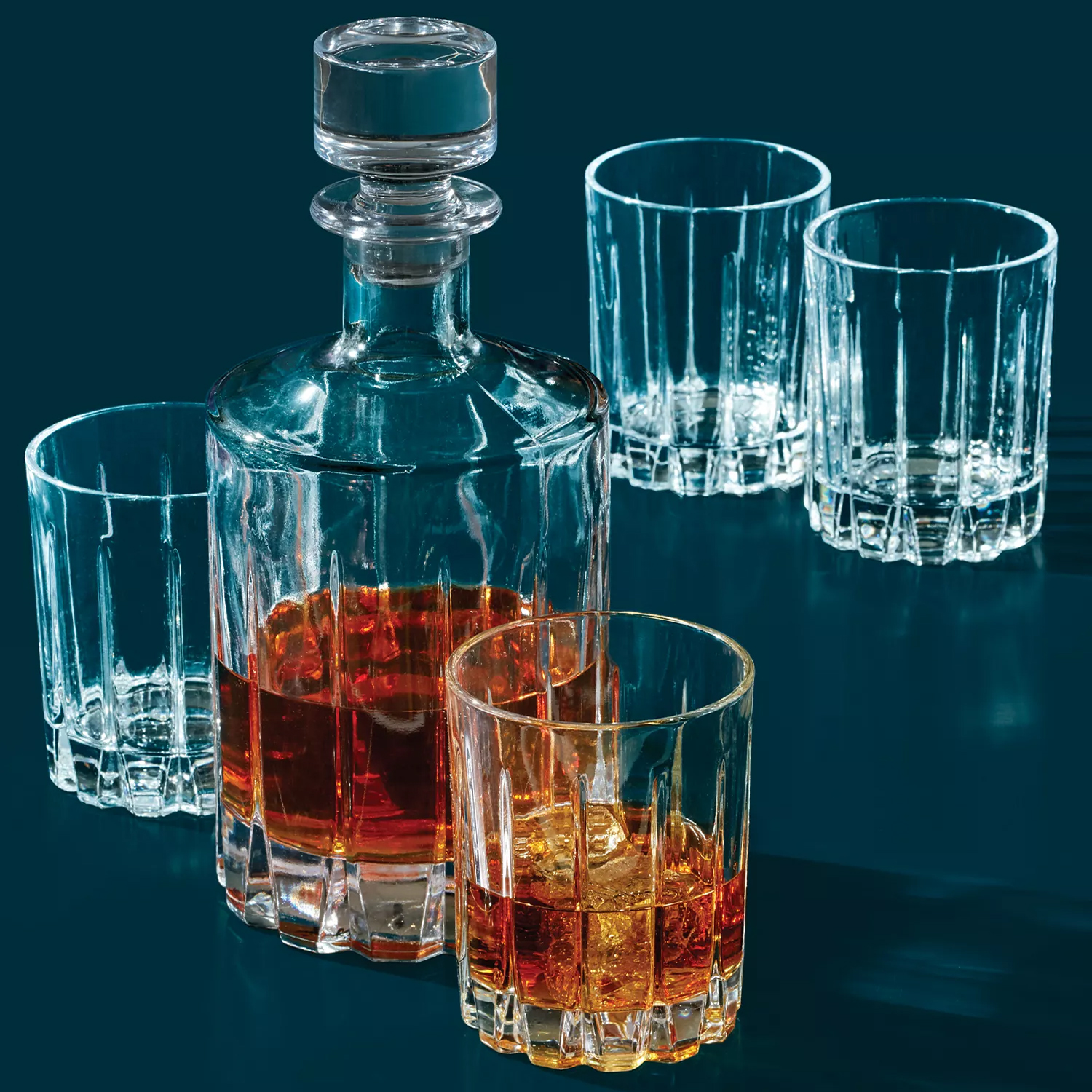 A clear glass decanter filled with amber liquid is surrounded by four empty matching glasses, one of which contains ice and the same liquid.