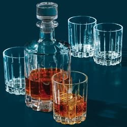 A clear glass decanter filled with amber liquid is surrounded by four empty matching glasses, one of which contains ice and the same liquid.