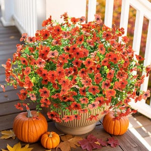 A pot of vibrant orange flowers is surrounded by three pumpkins on a wooden porch. Colorful autumn leaves are scattered around.