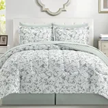 A bed is adorned with a floral-patterned comforter set, including matching pillow shams. The bedding features a white background with green leafy designs, complemented by light green accents.