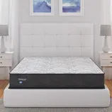 A Sleepys mattress is displayed on a white upholstered bed frame in a modern bedroom setup, flanked by white nightstands with matching lamps and abstract blue artwork above.