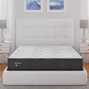 A Sleepys mattress is displayed on a white upholstered bed frame in a modern bedroom setup, flanked by white nightstands with matching lamps and abstract blue artwork above.