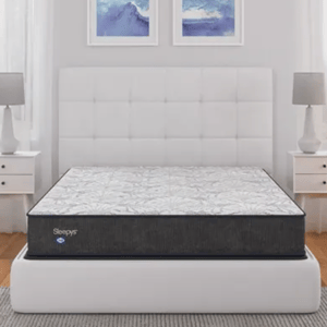 A Sleepys mattress is displayed on a white upholstered bed frame in a modern bedroom setup, flanked by white nightstands with matching lamps and abstract blue artwork above.