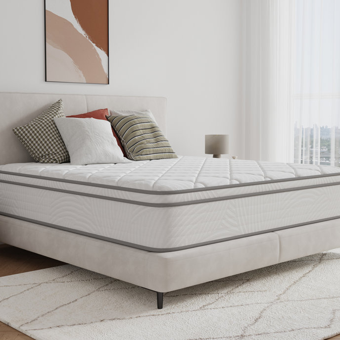 A 14-inch hybrid queen mattress with a quilted top, featuring a white and gray design. It rests on a light upholstered bed frame with a neutral-toned, modern bedroom setting, including decorative pillows and a patterned area rug.