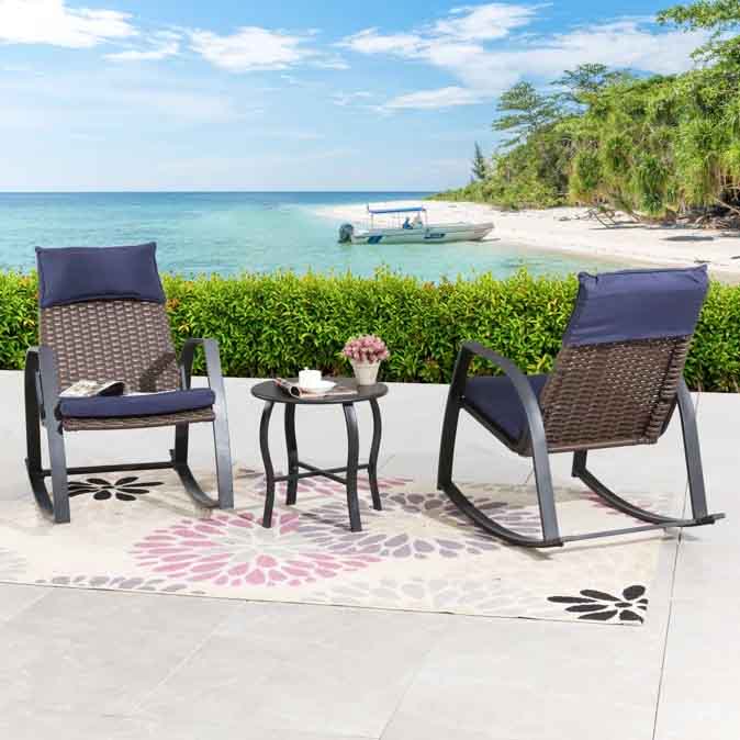 A 3-piece set featuring two wicker rocking chairs with navy cushions and a small round side table, set on a floral-patterned outdoor rug, overlooking a scenic beach view.