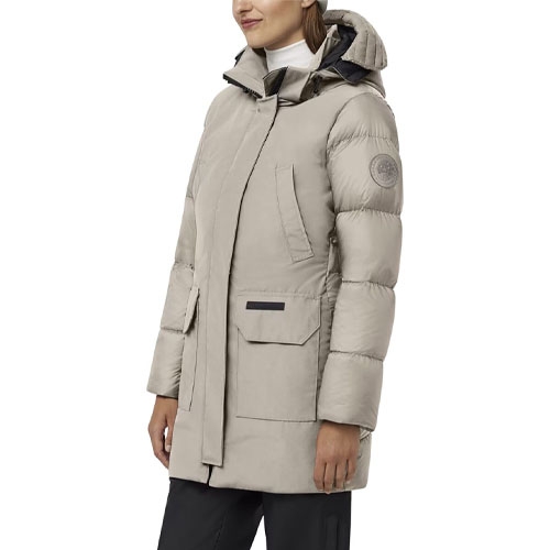 A woman is wearing a light gray down jacket with a hood and long sleeves, featuring a logo patch on the upper sleeve and large front pockets.