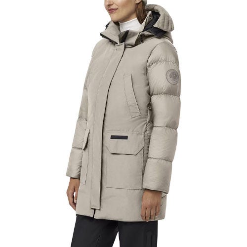 A woman is wearing a light gray down jacket with a hood and long sleeves, featuring a logo patch on the upper sleeve and large front pockets.