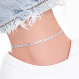 A silver anklet with an intricate pattern is worn on an ankle, paired with frayed hem jeans and a white sneaker.