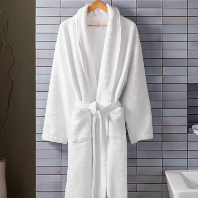 A white, plush cotton spa robe with a shawl collar, waist tie, and two front pockets, hanging on a wooden hanger against a tiled background.
