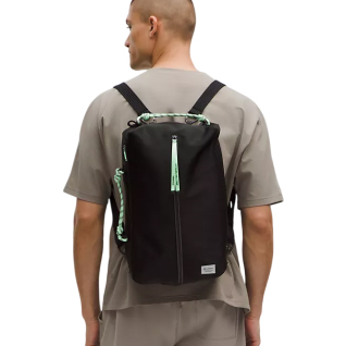 A man wearing a lululemon 16L Paracord Backpack, featuring a sleek black design with mint green paracord details, adjustable straps, and a top zipper closure. It's worn against a neutral-toned outfit.