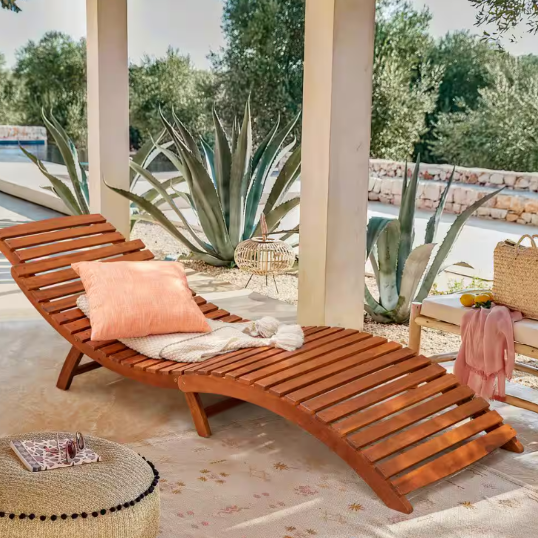 Wooden outdoor lounger with an orange pillow and blanket, set on a patio beside agave plants. Nearby, a small table with a basket and lemonade, and a pouf with sunglasses and a book on top.