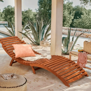 Wooden outdoor lounger with an orange pillow and blanket, set on a patio beside agave plants. Nearby, a small table with a basket and lemonade, and a pouf with sunglasses and a book on top.