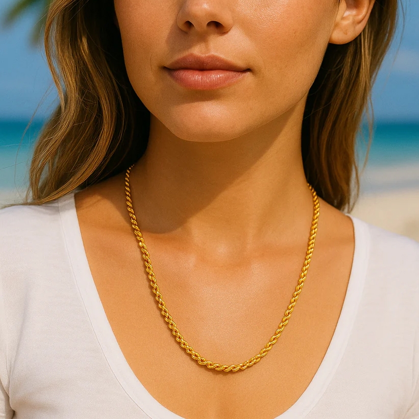 A gold rope chain necklace is worn by a person in a white shirt, set against a beach background.