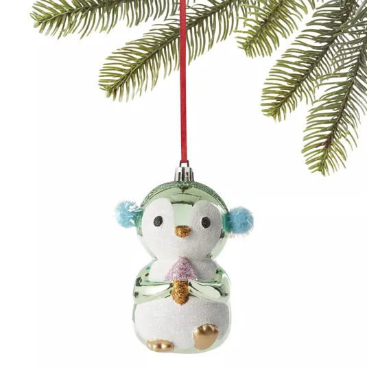 Glass penguin ornament with a glittery body, gold feet and beak, blue earmuffs, and a pink scarf, hanging from a red string beneath tree branches.