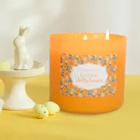 An orange candle labeled \“Colonial Candle Lemon Jellybean\“ is next to a white ceramic bunny on a stand, surrounded by small yellow-speckled eggs.