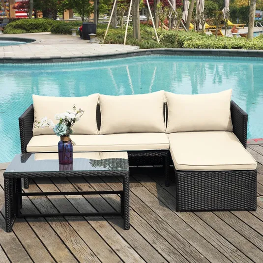 Outdoor patio set with a wicker sectional sofa featuring cream cushions and a glass-topped coffee table; set is placed on a wooden deck by a poolside, decorated with a floral arrangement in a blue vase.