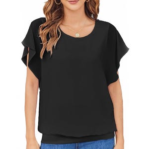 A woman is wearing a black batwing top with loose, flowing sleeves and a round neckline, paired with blue jeans. She has wavy, light brown hair and is accessorized with a round pendant necklace and hoop earrings.