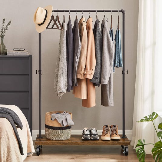 Clothing rack with jackets, shirts, and hats, featuring a bottom shelf holding a woven basket and two pairs of shoes. A dresser, a plant, and a bed appear nearby.
