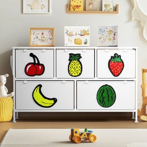 A storage cabinet with six drawers features embroidered fruit designs: cherries, pineapple, strawberry, banana, and watermelon. The cabinet is set in a child's room with framed illustrations and toys around it.