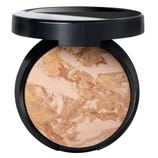 A compact powder with a marbled blend of beige, gold, and light brown shades, encased in a black container with a flip-open lid.