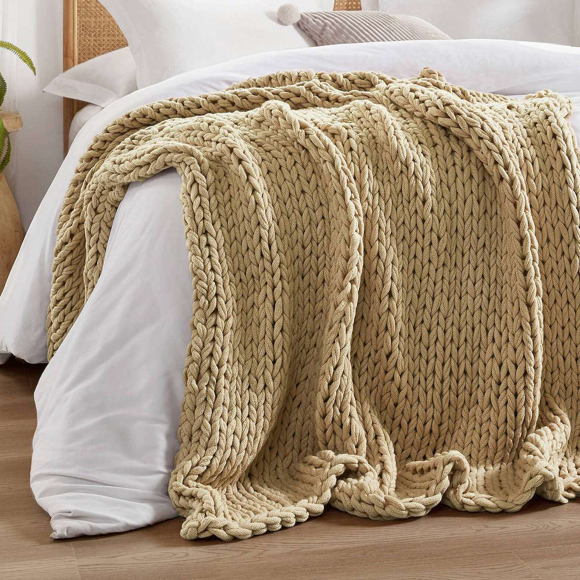 Madison Park Chunky Handmade Throw features a thick, woven texture in a neutral beige color, draped over a bed. The knit style is intricate and voluminous, adding warmth and style to the setting.