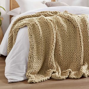 Madison Park Chunky Handmade Throw features a thick, woven texture in a neutral beige color, draped over a bed. The knit style is intricate and voluminous, adding warmth and style to the setting.