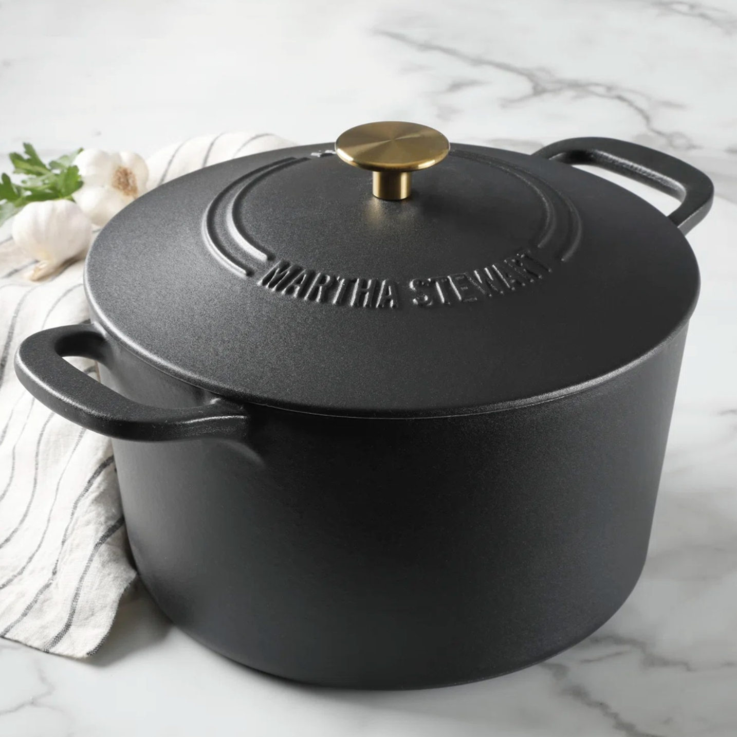 A black cast iron Dutch oven with a brass handle on the lid, featuring the name \“Martha Stewart\“ embossed on top.