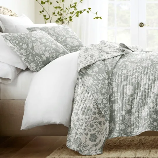 A bed is made with a floral-patterned quilt and matching pillows, accompanied by white sheets. A wooden nightstand with a lamp and plant completes the cozy bedroom setting.