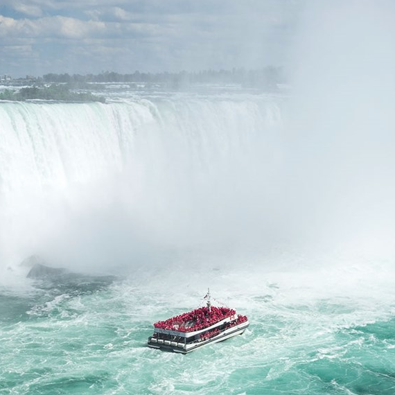 A tour boat filled with people in red ponchos approaches the misty base of Niagara Falls against a backdrop of powerful cascading water.